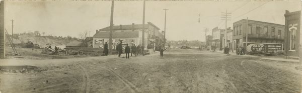 Panoramic street view, with small groups of people scattered about, and standing on the sidewalks near buildings lining the unpaved street. On the sides of many of the buildings are painted advertisements or signs such as: "Badger State Banner," "First National Bank," "Cole's Cough Cure(?) Cures a Cough. Children Like it- So will (?)" and "City Bakery". In the background is the river, and houses on a hill on the other side.