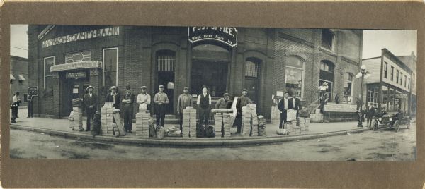 Panoramic view from street of sixteen men standing on the sidewalk in front of the Jackson County Bank and the Post Office. On the sidewalk in front of the men are large stacks of paper, and there is a parked automobile on the right. Text on the back of the print of this image reads: "South side of Main — corner shown is 1st St." Behind the men in front of the Post Office is a sandwich board with a poster that reads, in part: "Fighting," Great War...," and "Ask the Postmaster."