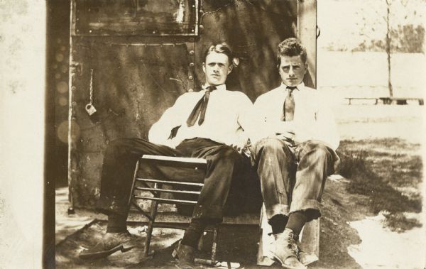 Outdoor portrait of two young men, sitting and leaning the backs of their chairs against a wall or door. The young man on the left is looking straight ahead, while the young man on the right (identified as Harold Van Schaick), is looking off to the side. Both men are wearing dark pants, white shirts, and ties.