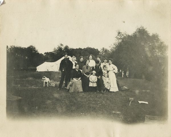 A group of thirteen men, women, and children posing outdoors for a group portrait. A dog is standing off to the left. According to text on the back of the print, some individuals can be identified as: "left to right, standing- 1. 2. Mrs. Wayland Cole 3. Julia Bright 4. Kathryn Cole 5. 6. 7. Birdine Perry Jones- sitting- 1. 2. Florence O'Hearn 3. - and three children Willard Jones, Dorothy Krohn." In the background there appears to be a tent set up amid some trees.