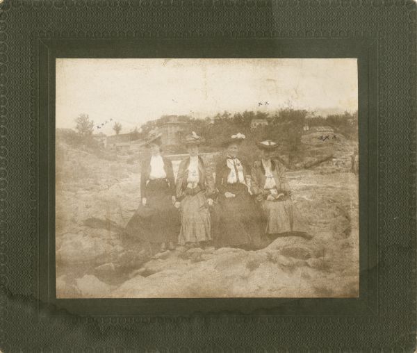 A group of four women posing on river rocks. Each woman is wearing a long skirt, blouse, and hat. The first woman on the left is identified as being Tillie Walden Johnson. The scenery behind them includes the Spaulding Wagon Shop, the Old Boat House, Old Weber Residence, and the Old Spaulding Boarding House.