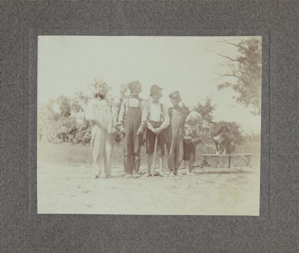 Group portrait of five young boys and a dog, posing standing in a line in front of some trees. The dog is at the end of the line on the right, sitting on top of a wooden wagon/cart. All the boys are wearing a variation of a shirt and overalls, and all boys except for the youngest one on the very right is wearing a hat. According to text on the back of the print of this image, the first boy on the left is identified as "Hans Hanson (Judge)," and the boy in the center is "Alfred Brandon."