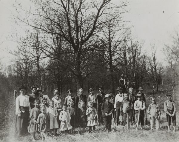 A large group of both boys and girls posing standing outside. Text on the back of the print of this image reads: "South Alma, Wis. About 1900. School pupils with their teacher (extreme left), Miss Nellie Adams. This school was located in Jackson County, on the site now occupied by a cheese factory."