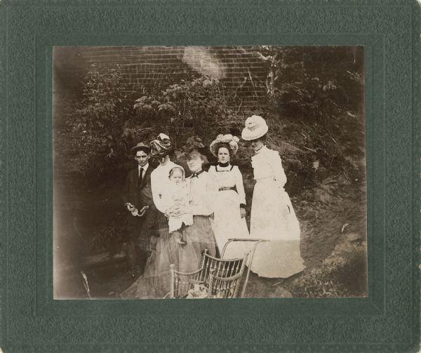 Group portrait of four women, one man, and one infant standing outdoors in front of a brick wall covered in vines. The man is on the far left next to one woman, and the woman in the center is holding the infant. It looks as if another individual is crouching behind the woman holding the infant, which may be a partial double exposure or blurring. The other two women are standing on the right. Each woman is wearing an elaborate hat, and there is either a chair or a stroller in the foreground.
