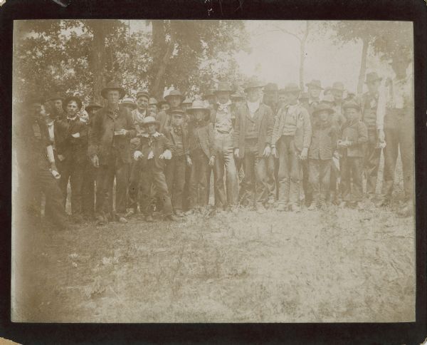 A group of many men and a few boys, standing in a clearing surrounded by trees. Text on the back of the print of this image identified them as, "men of the same crowd."