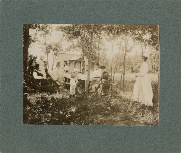 A group of women and children sitting and standing around a long table in a grove of trees enjoying a picnic. A man is sitting on a couch on the left, and a woman in a white dress is standing off to the right. A white house is in the background.