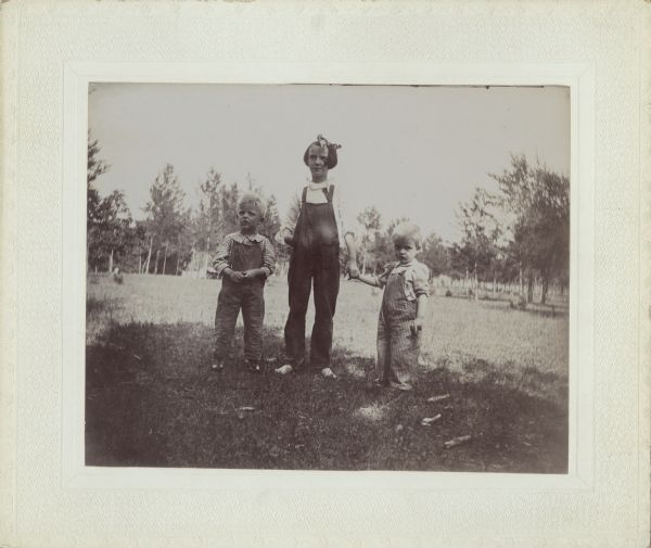 A portrait of three children posing standing outdoors in a field. Each child is wearing a shirt and overalls. The older girl standing in the center is holding the hand of each one of the children.
