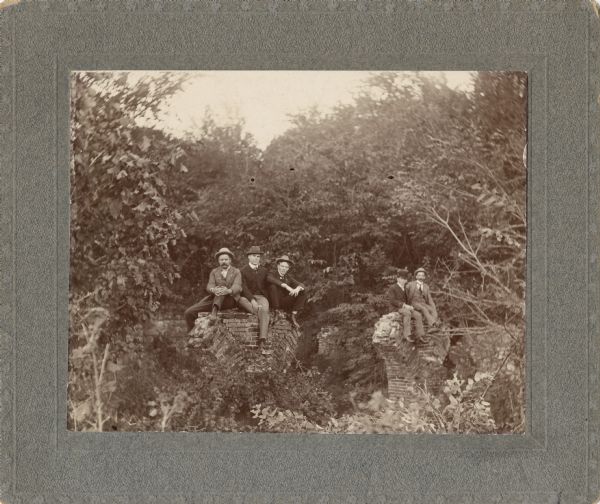 Group portrait of five men posing sitting on top of brick ruins, perhaps the remains of bridge supports.
