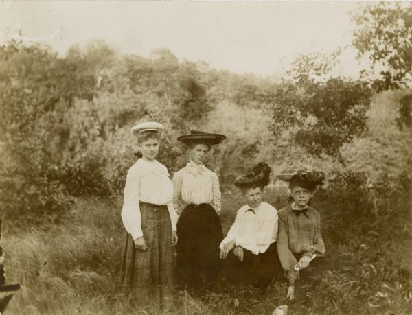 Outdoor group portrait of four women, taken in a field. The two women on the left are posing standing, while the two on the right are posed kneeling or crouching. The women are wearing skirts, blouses and hats.