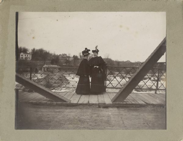 View from road of two women posing standing on a bridge. Both women are wearing black skirts/dresses and capes along with elaborate hats. Behind them is the rushing river, as well as several buildings on the riverbank. One building is painted on both the roof and side with advertising which says: "Carbollsalve."