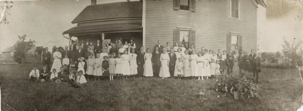 Panoramic view of a large group of people posing standing in front of a house, gathering for a wedding. The bride and groom are standing in the center.