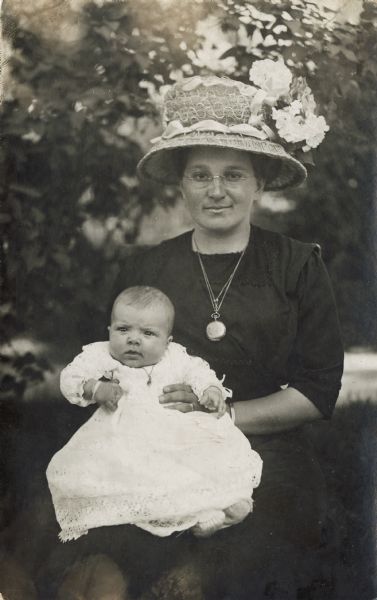 Outdoor portrait of a woman sitting and holding an infant on her lap. The woman is wearing a dark dress, glasses, a large hat with flowers on it, and silver jewelry including a large locket. The infant is wearing a white dress and a necklace.
