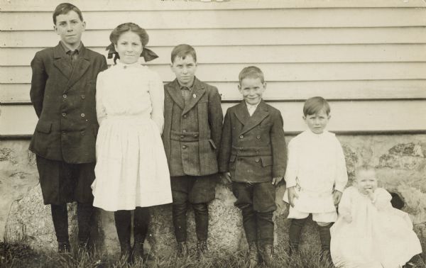 Outdoor group portrait of five children, posing standing against the side of a building. They are standing in a line in descending height order, presumably with the oldest boy on the left, and the youngest on the right. There are four boys, one girl, and one infant.
