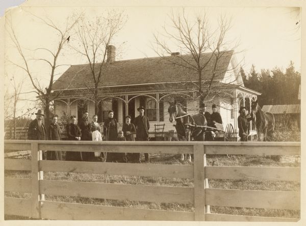 An outdoor group portrait of twelve people and two horses. The four men, seven women, one child, and two horses are all standing in front of a house. There is a fence in the foregrund. On the right, one man is holding the reins of one horse, while the other horse is hitched to a carriage which is carrying a man and a woman. The rest of the people are posing in a group on the left.