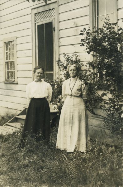 Portrait of two older women posing standing in front of a house near steps leading up to a door. Both women are wearing blouses and long skirts, and have their hair pulled back. The woman on the right is also wearing a long necklace.
