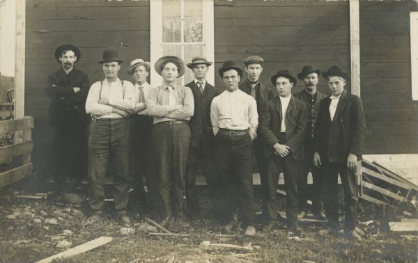 Outdoor portrait of ten men posing standing in front of a building. All of the men are wearing hats, and some are wearing ties.