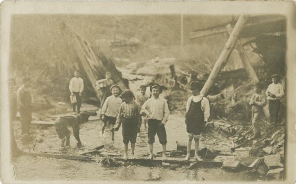 A view across water towards a group of boys playing in and around the water near a shoreline. Many of the boys are barefoot. They are surrounded by wreckage of some sort, including broken rocks, fallen timber, etc.