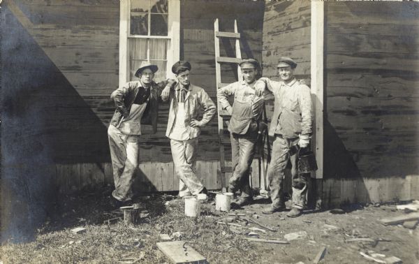 A portrait of four men posing standing in two pairs in front of a building. Each pair of men have their arms around each others shoulders, and all the men are wearing overalls and caps. Cans of paint and paintbrushes are sitting on the ground in front of the men, and the man on the right is holding both a paintbrush and can of paint in his left hand.