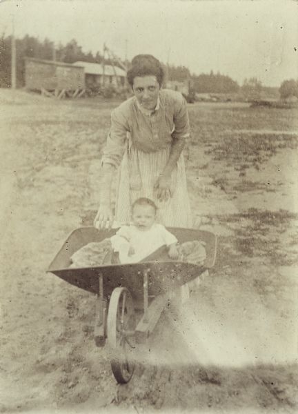 A view of a woman feeding an infant with a bottle while outdoors. The infant is sitting in a wheelbarrow in front of her. A scattering of buildings are in the background.