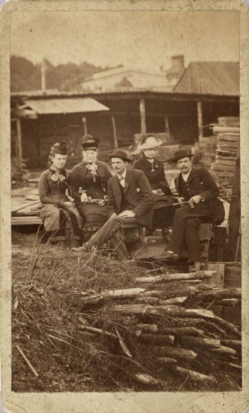 Outdoor portrait of three women and two men, all posing sitting on a wooden platform, perhaps in a lumber yard. A large open shed structure and stacks of timber are in the background. The women are wearing dresses and hats, and the men are wearing suits, ties and hats.