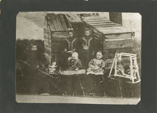 Outdoor portrait of five children and various dolls in front of a small wooden structure or cabin. A boy is standing on the left side, holding the handle to a wagon in which an infant is sitting. A girl is sitting in a chair next to the wagon on the right, and to the left of the wagon is a small stroller holding a doll. Two girls standing behind the wagon are holding pots and pans. On the right two dolls are sitting in a small toy lawn swing.