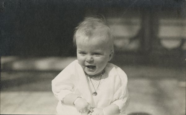 Outdoor portrait of a laughing baby wearing a white shirt/dress and a locket necklace.