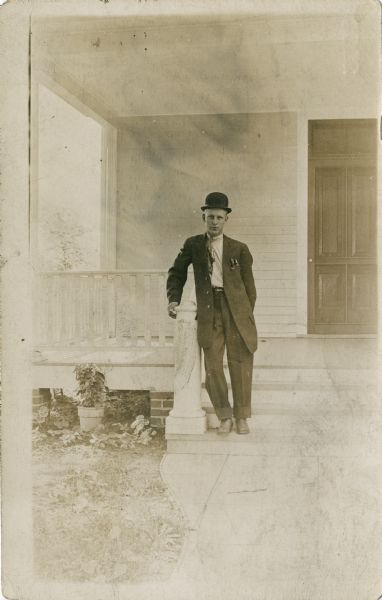 Outdoor portrait of a man standing on steps leading to the porch of a house. He is leaning on the railing and is wearing a suit, tie and hat.