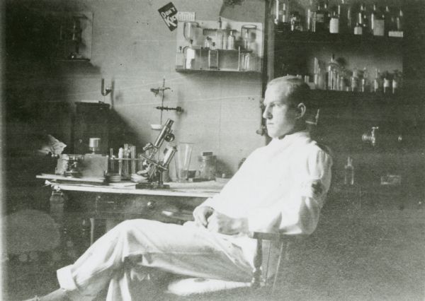A view of a male medical student sitting on a chair facing toward the left. Behind him is a desk, and shelves full of instruments and bottles. A microscope is on the desk. Text on the back of the print of this image reads: "Early 20th century, from Black River Falls, Wis. From Jackson County Historical Society. Copy neg loaned by UW Dept. of History of Medicine, 1977." This image was probably captured by photographer Charles Van Schaick.