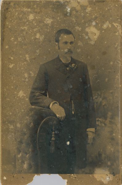 Three-quarter length portrait of a man posing standing and resting his right arm on a curved-back chair. He is wearing a suit and tie, and has a flower in his lapel.