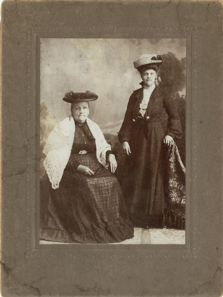 Full-length studio portrait of two women posing in front of a painted backdrop. The woman sitting on the left is older and is wearing a white shawl over her shoulders. The woman on the right is standing, and has her left hand resting on the back of a cloth covered chair. Both women are wearing elaborate hats.
