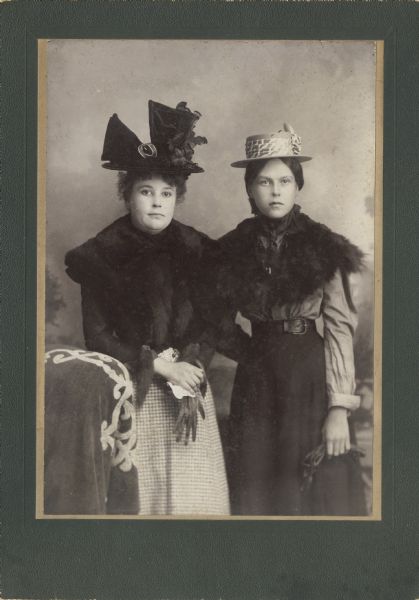 Three-quarter length studio portrait of two women posing standing in front of a painted backdrop. Both women are wearing elaborate hats and fur shawls, and are holding gloves in their hands.