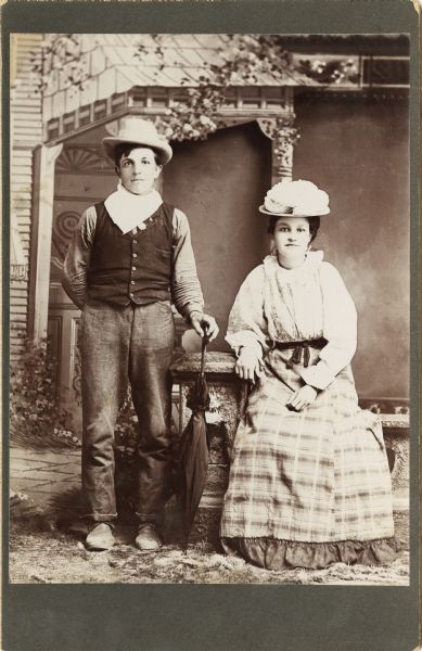 Full-length studio portrait of two youth posing in front of a painted backdrop with a prop stone wall. The young man on the left is standing and holding an umbrella in his left hand, and leaning against the prop stone wall. He is wearing a vest, hat and a bandana around his neck. The young woman is on the right sitting on the prop stone wall. She is wearing a skirt, blouse, and a large hat.