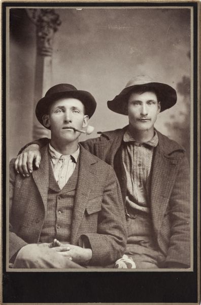 Waist-up studio portrait of two young men posing sitting in front of a painted backdrop. They are wearing unbuttoned coats, and hats. The young man on the right has a moustache and has his arm around the other man's shoulder. The young man on the left has a pipe in his mouth.