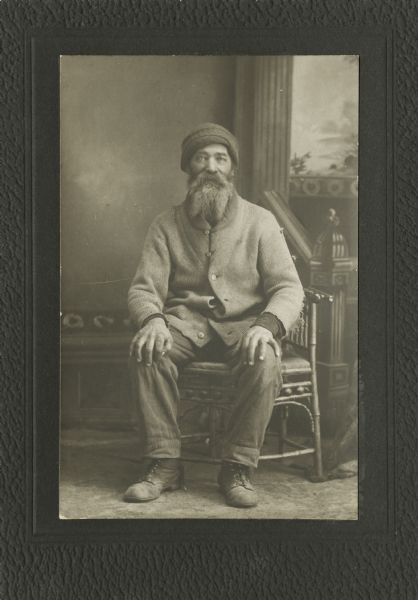 Full-length studio portrait of a man sitting in front of a painted backdrop. He has a beard and moustache, and is wearing a hat and a somewhat tattered coat.