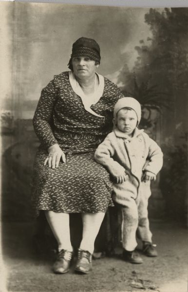 Full-length studio portrait of a woman sitting next to a standing child in front of a painted backdrop. The woman is wearing a printed dress and cloche hat. On the right, the child is wearing a jacket (possibly a snowsuit of some sort) and a hat.