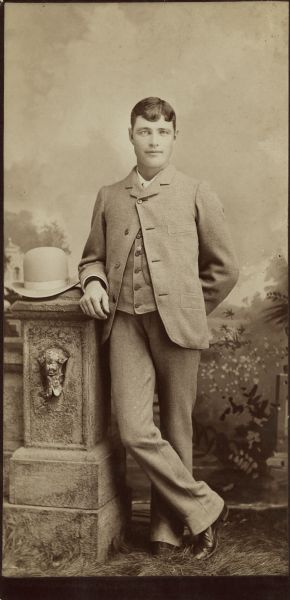 Full-length studio portrait of a young man standing in front of a painted backdrop and resting his right arm on a prop stone wall. He is wearing a suit and suit coat, and has his hair parted. His hat is resting on top of the stone wall.