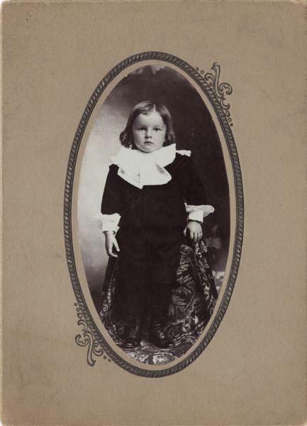 Full-length oval framed studio portrait of a child posing standing on a covered chair. The child is wearing a dark-colored suit, and a shirt with white ruffles at the neck and sleeves.