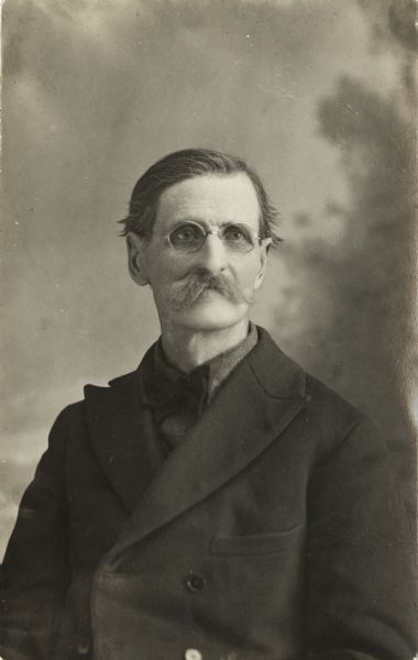 Waist-up studio portrait of an older, mustachioed man who is posing sitting in front of a painted backdrop. He is wearing a tie, coat and eyeglasses.