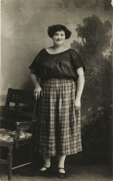 Full-length studio portrait of a woman standing in front of a painted backdrop next to a chair. She is wearing a blouse and skirt, and has a short hairstyle.