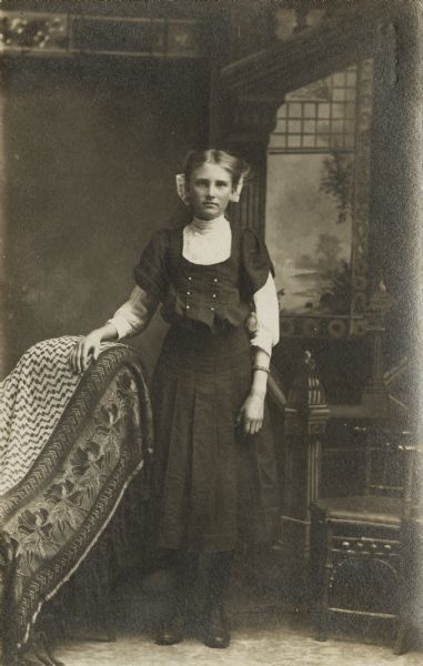 Full-length studio portrait of a young woman standing in front of a painted backdrop. She is resting her right arm on a covered chair and is wearing a dress, blouse and tall boots. Her hair is pulled back and she is wearing a large bow.