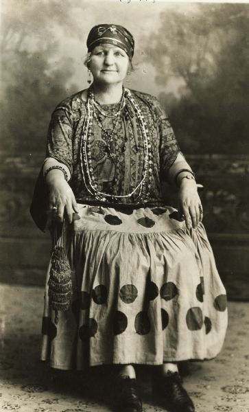 Full-length studio portrait of Emma W. Gebhardt sitting in front of a painted backdrop. She is wearing a long, polka-dotted skirt, a printed blouse, a bandana over her hair, earrings, and many necklaces and bracelets. She is also holding a small purse/satchel which is hanging from her right hand.<p>A series of portraits taken of guests at an Eastern Star "Gypsy Party".