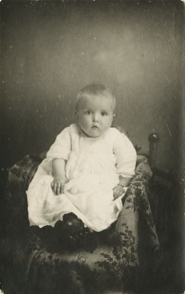 Full-length studio portrait of an infant sitting on a cloth covered chair wearing a white dress.
