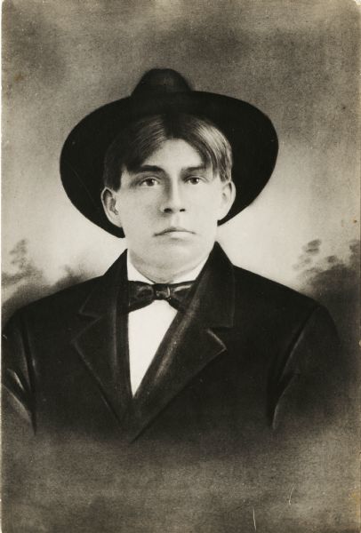 A heavily retouched waist-up studio portrait of a young man in front of a painted backdrop. He is wearing a suit coat and tie along with a hat.