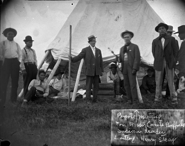 Outdoor portrait of many men in front of and near a tent, including Tom Roddy and Henry Stacy. Tom Roddy, "known as White Buffalo," is standing to the right of the man who is also standing and holding a large pole/stick. Henry Stacy is posed sitting to Tom Roddy's left, and is additionally identified as being the husband of Mountain Wolf Woman. This image was possibly taken at a peyote meeting.
