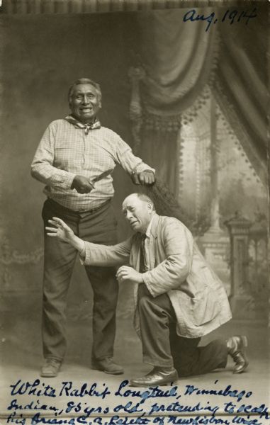 Full-length studio portrait of an 85-year-old Ho-Chunk man, White Rabbit Longtail, posing standing and smiling while pretending to scalp his friend, C.A. Leicht of New Lisbon, who is posing on one knee with outstretched arms and a pretend grimace.