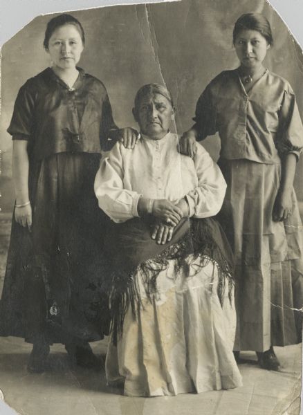 Full-length portrait of two Ho-Chunk women standing on either side of an older Ho-Chunk woman who is sitting. Each woman is wearing a long skirt, a blouse, and jewelry, and the woman sitting also has a shawl on her lap. Both of the standing women are resting a hand on the seated woman's shoulders.