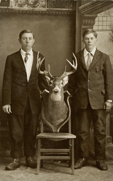 Two young men, Carl and Roy Linnell, standing on either side of a wooden chair supporting a mounted buck's head with antlers. They are wearing dark suit jackets with neckties, and boots with their trousers rolled up over them. They are standing in front of a painted backdrop.