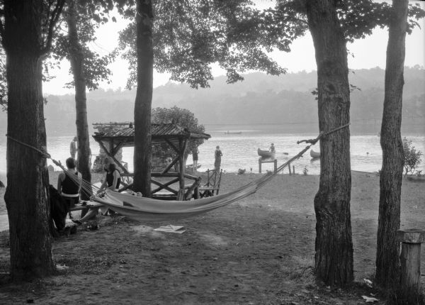 A group of people picnics near a hammock hanging between two trees along a beach.  Several people canoe in the water.