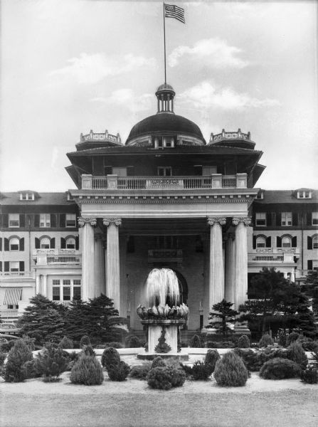 View of the Monument Hotel front entrance, featuring Greek columns, decorative foliage, and a fountain. An American flag flies from atop the building's cupola.