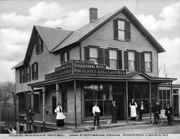 View from street towards men, women, and a dog or puppy, standing along the wrap-around porch of the Susquehanna Hotel. The street or yard on the left of the hotel drops steeply, and a lower level of the building can be seen. The signs on the porch's roof read: "John S. Nittrowr," "Susquehanna Hotel," and "Hinchliffe's Ales & Lager Beer."